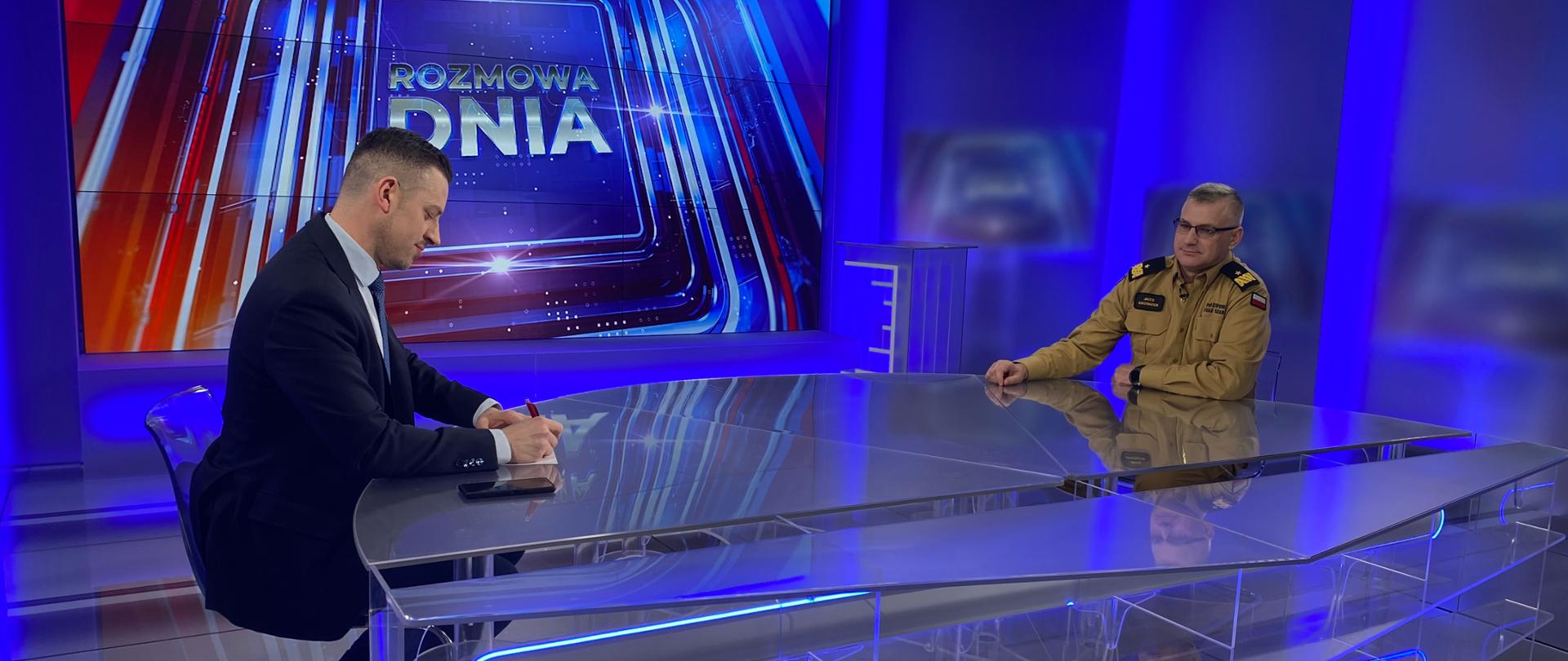 W studio telewizyjnym, przy stole siedzi redaktor i komendant wojewódzki PSP w mundurze generalskim, służbowym. Stół przeźroczysty, w tle wystrój wystrój studio.
