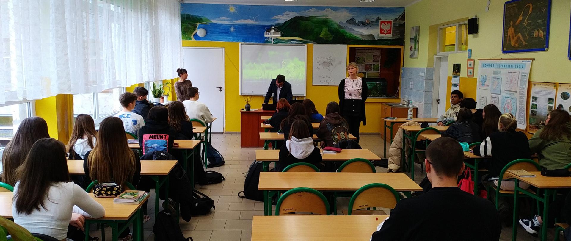 W klasie w ławkach siedzi grupa kilkunastu dziewczyn i chłopców. Są odwróceni plecami. Na drugim planie przy tablicy stoją dwie kobiety i mężczyzna.