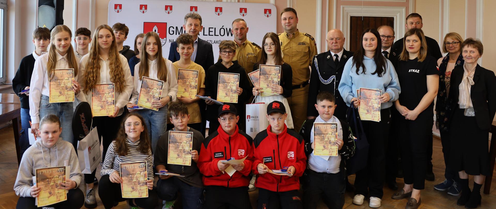 Na zdjęciu w sali widać komisję, składającą się z dwóch strażaków reprezentujących Komendę Miejską Państwowej Straży Pożarnej w Częstochowie, w umundurowaniu służbowym, w składzie: młodszy brygadier Przemysław Klita oraz młodszy aspirant Adrian Nowak, Wójt Gminy Lelów – Pan Krzysztof Molenda, Prezes Zarządu Gminnego Ochotniczej Straży Pożarnej druh Władysław Jaworski oraz Komendant Gminny Ochotniczej Straży Pożarnej druh Tomasz Świątek. Przed komisją stoją laureaci z grupy wiekowej klas 5 do 8 szkoły podstawowej wraz z ich opiekunami. W tle można dostrzec biały baner promujący gminy Lelów, na którym znajduje się herb miejscowości, przedstawiający wieżę kościoła z murem na czerwonym tle. Cała sceneria przedstawia uroczystość wręczenia nagród za osiągnięcia dzieci w dziedzinie bezpieczeństwa i działalności strażackiej. Atmosfera wypełniona jest radością z otrzymanych nagród.