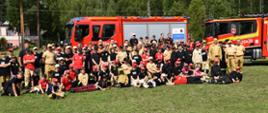 Zdjęcie plenerowe uczestników obozu na tle dwóch czerwonych samochodów strażackich ustawionych na trawniku. W tle wysokie zielone drzewa. 