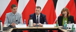 Za stołem siedzą minister Czarnek, wiceminister Piontkowski i kobieta w czarnej marynarce, za nimi rząd polskich flag.