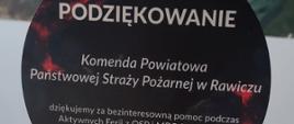 Na stoliku stoi statuetka w kształcie koła. Na niej znajduje się następujący napis: PODZIĘKOWANIE Komenda Powiatowa Państwowej Straży Pożarnej w Rawiczu dziękujemy za bezinteresowną pomoc podczas Aktywnych Feriiz OSP i MDP Sobiałkowo. Na dole znajduje sie logo MDP oraz OSP Sobiałkowo.