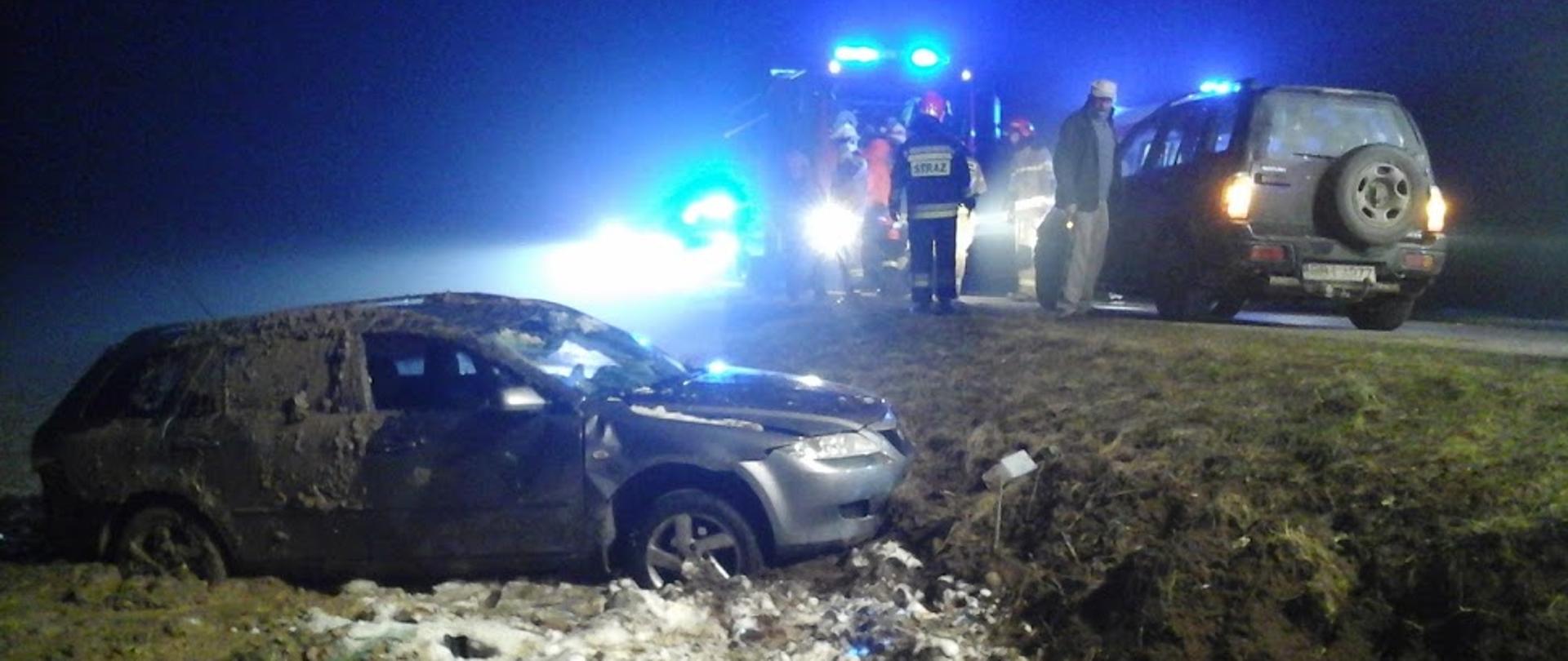 Na zdjęciu widać rozbite auto postawione na koła w przydrożnym rowie, w tle wozy ratowniczo-gaśnicze