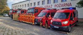 strażacy psp i osp stojący w szeregu przed samochodami pożarniczymi, za samochodami budynek jednostki ratowniczo-gaśniczej