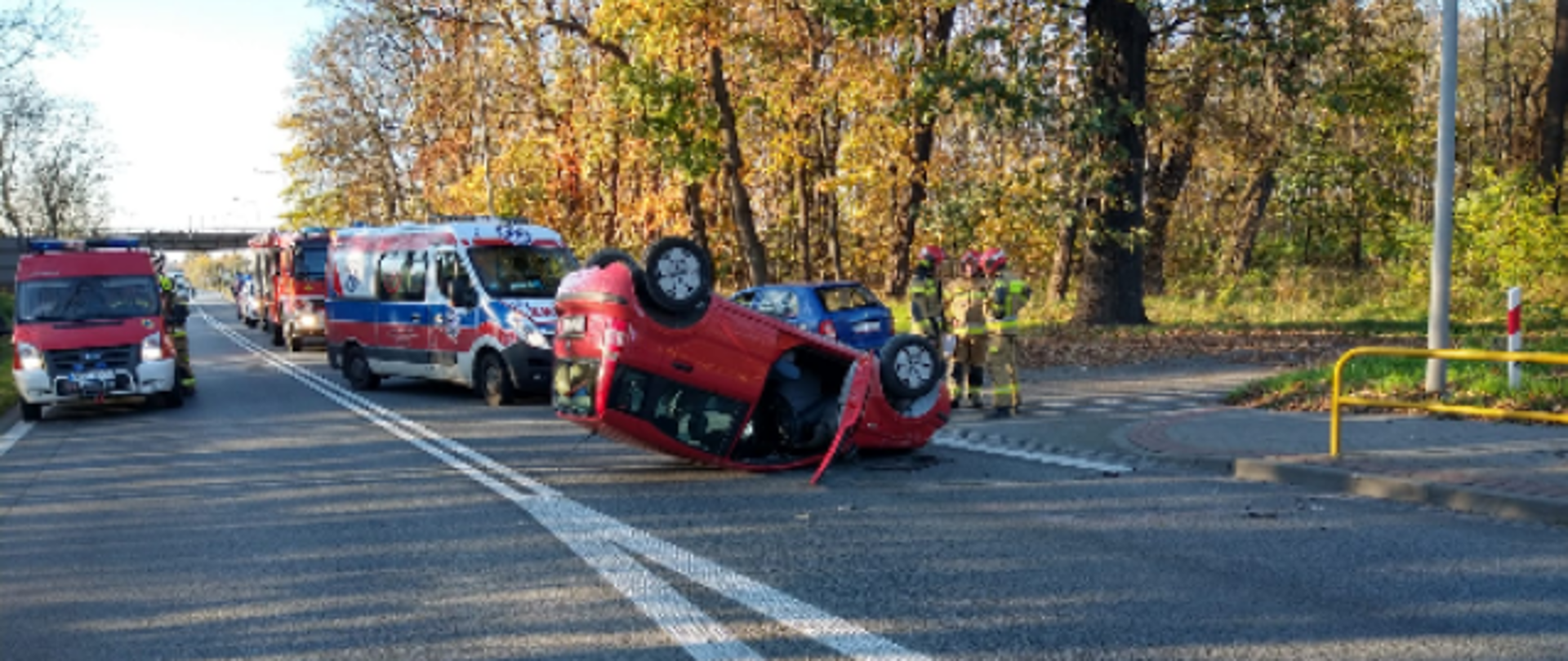 Na zdjęciu znajduje się samochód wywrócony na dach, w tle widać stojące samochody strażackie i karetkę pogotowia na drodze