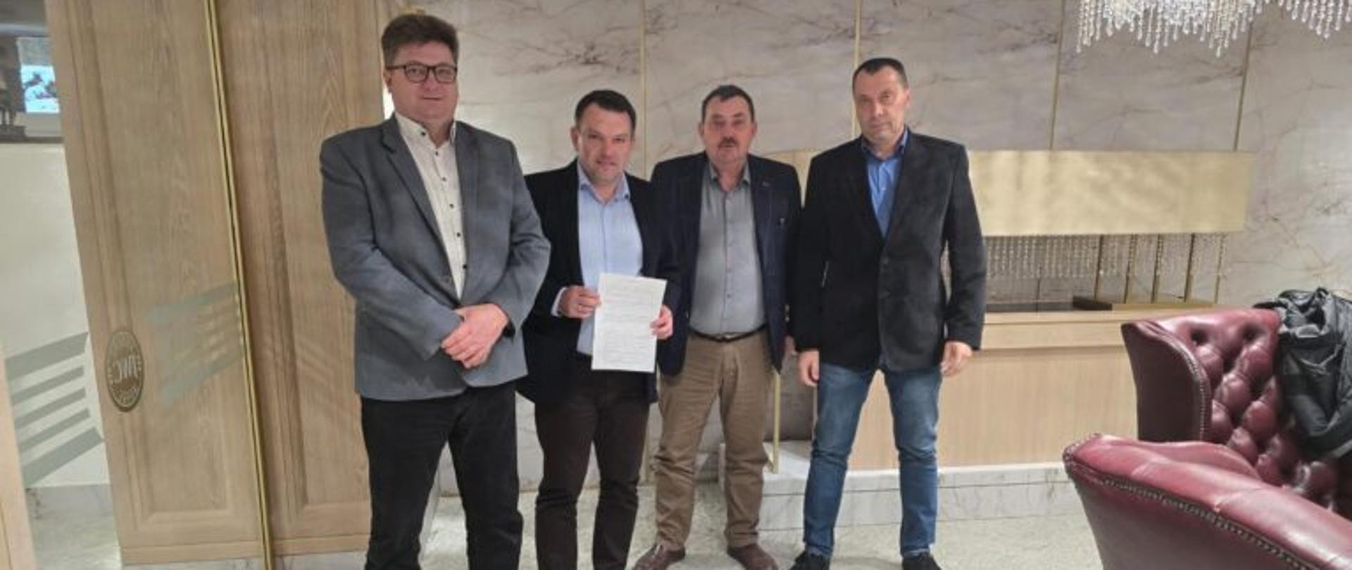 Czterech mężczyzn stoi w reprezentacyjnym wnętrzu. Jeden z nich trzyma w dłoniach dokument skierowany w stronę aparatu. Pozostali stoją obok, wszyscy zwróceni przodem do obiektywu.