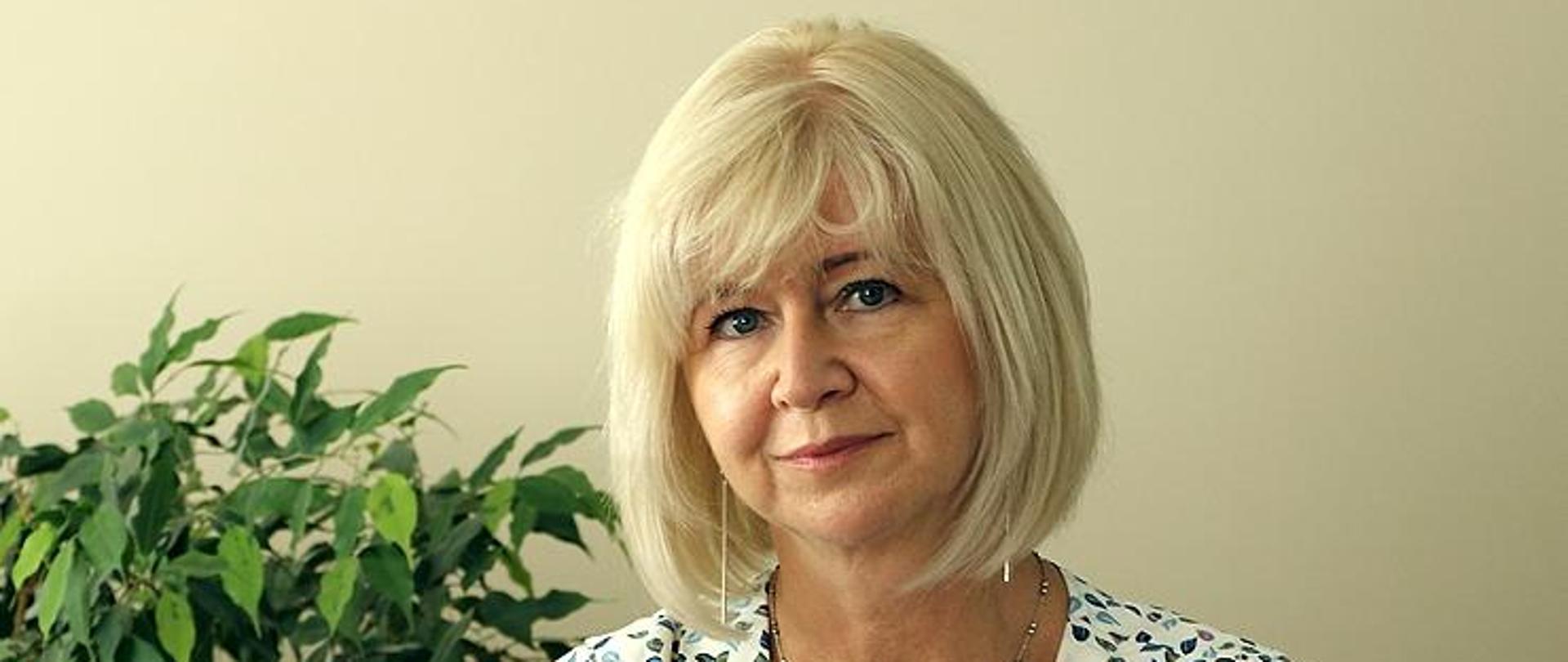 Wizerunek Regionalnego Dyrektora Ochrony Środowiska w Olsztynie. Kobieta siedzi przy stole, na którym leży otwarty dokument. W ręku trzyma długopis.