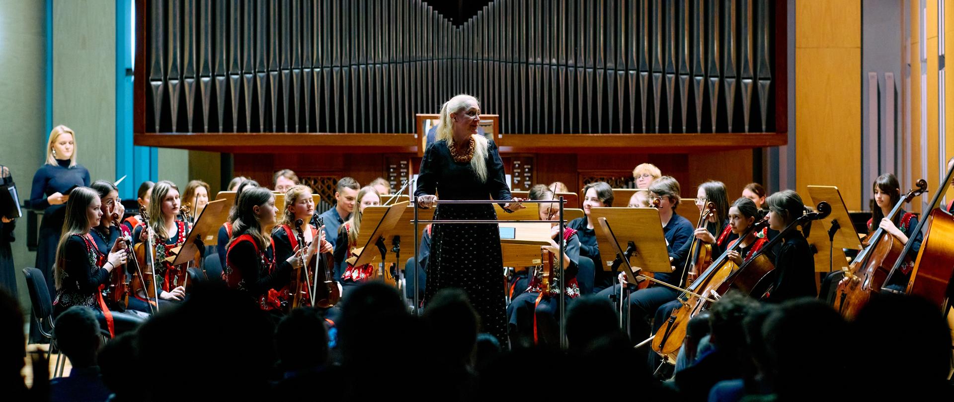 Na scenie sali koncertowej uczniowie orkiestry siedzą trzymając swoje instrumenty, przed nimi pulpity z nutami. Za nimi organy, z lewej strony zespół wokalny. Na pierwszym planie publiczność. Przed orkiestrą stoi dyrygentka skierowana do publiczności.