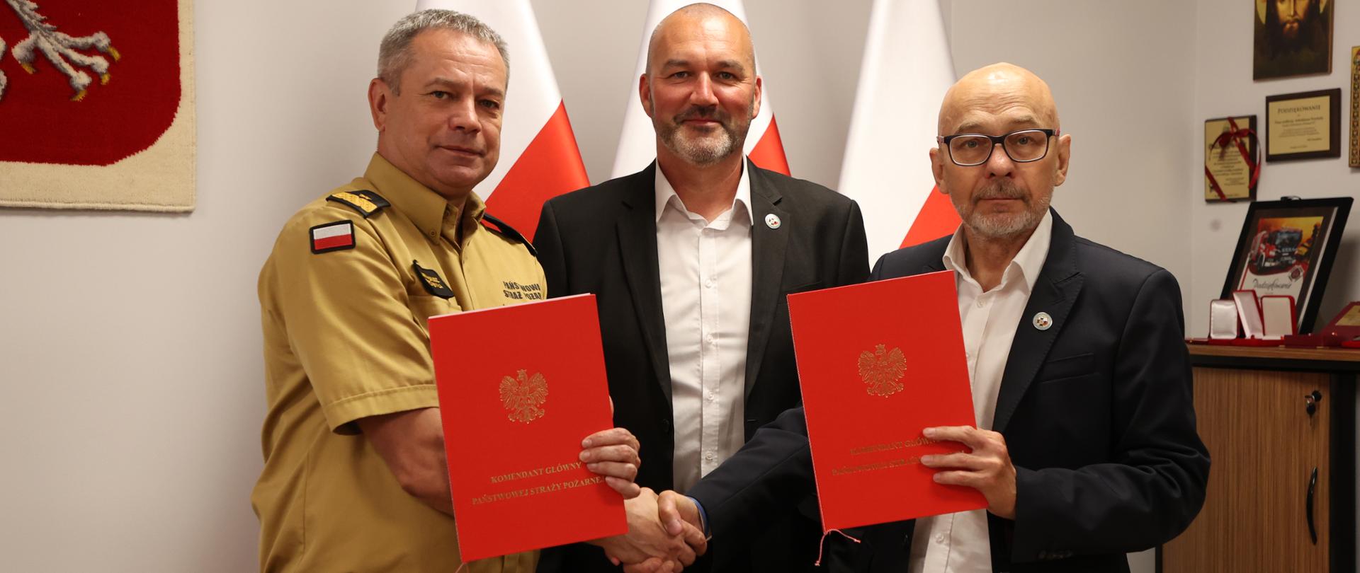 Zastępca komendanta głównego PSP (od lewej strony) w mundurze służbowym koloru piaskowego stoi wraz z dwoma mężczyznami w garniturach i prezentują dwie czerwone teczki zawierające podpisane porozumienie, za nimi 3 flagi RP