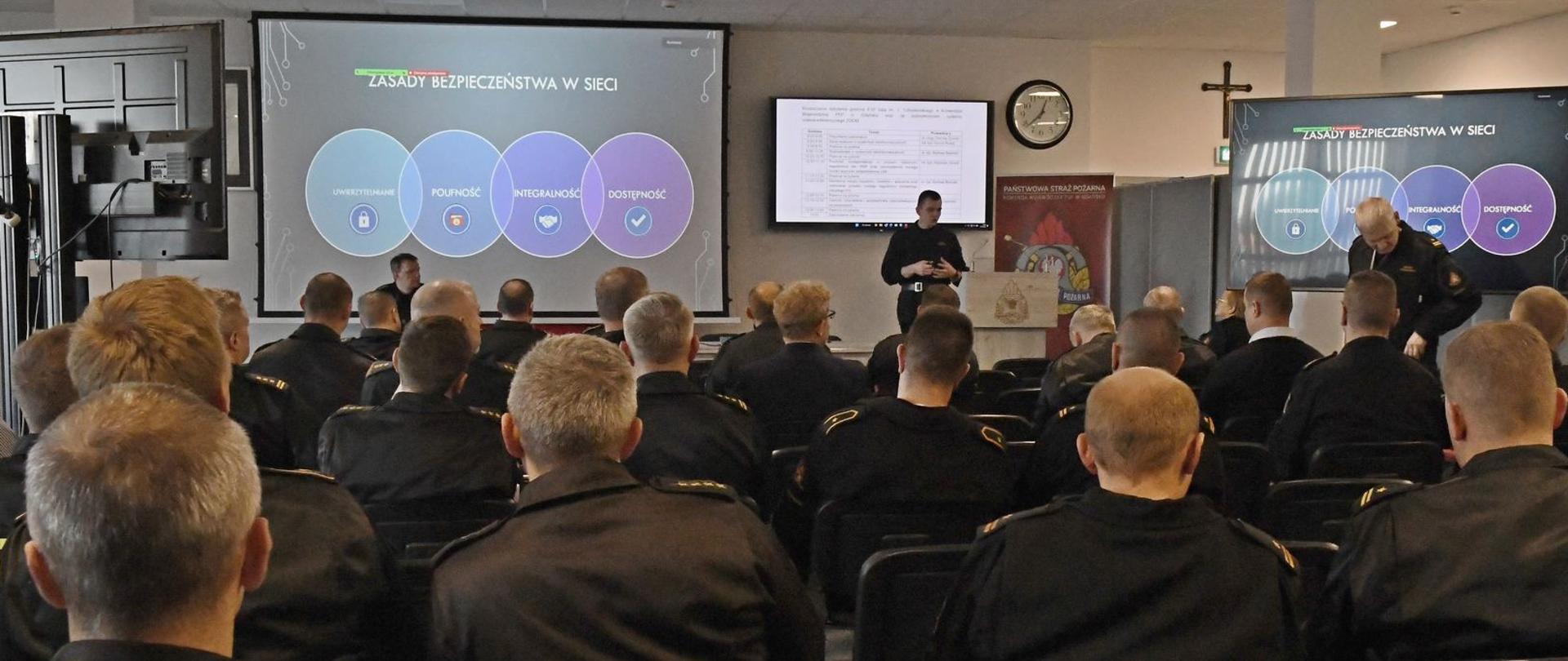 Zdjęcie przedstawia funkcjonariuszy uczestniczących w szkoleniu oraz funkcjonariusza Komendy Wojewódzkiej PSP prowadzącego spotkanie.. Na zdjęciu widać slajd z prezentacji dot. bezpieczeństwa w sieci.