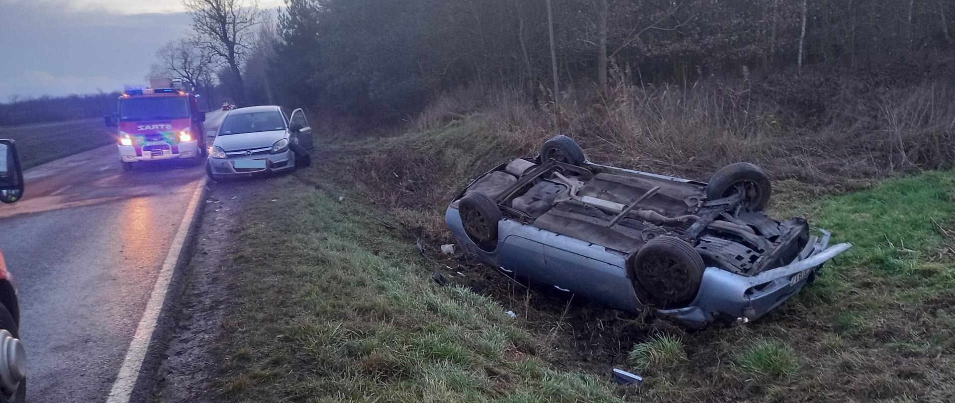 Widać samochód w pozycji na dachu w rowie, na poboczu jest drugi pojazd, za nim samochód strazacki