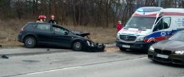 Zdjęcie przedstawia samochód Seat Leon z rozbitym przodem stojący częściowo na jezdni, a częściowo na chodniku. Z prawej strony widać karetkę Pogotowia Ratunkowego.