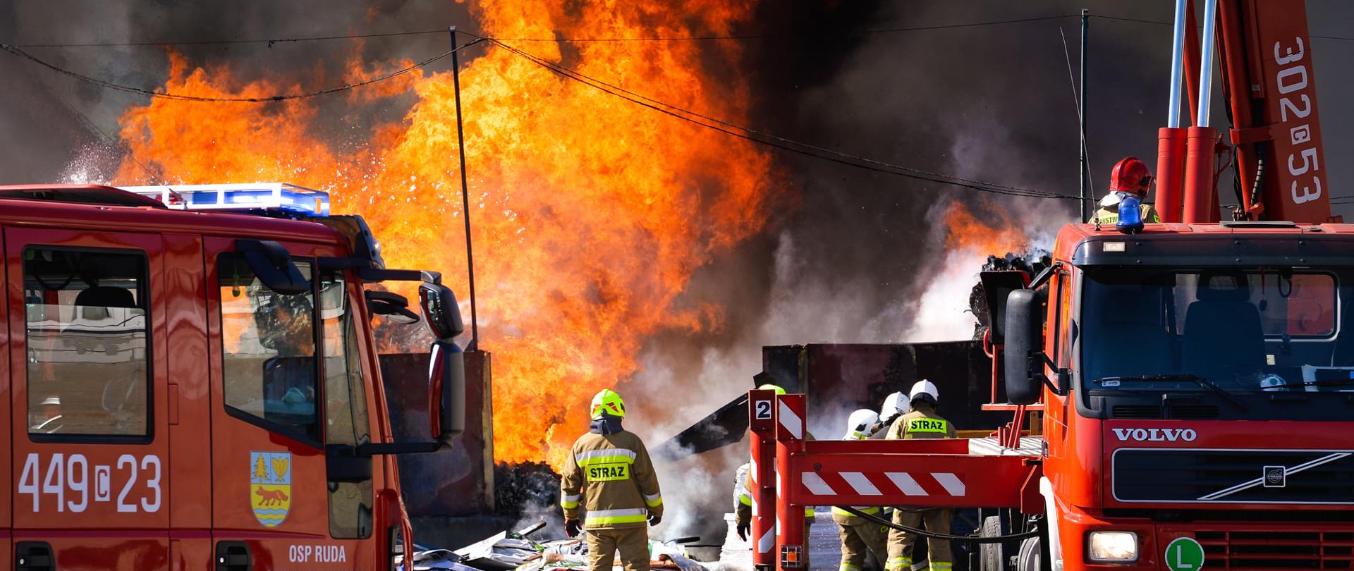 Na pierwszym planie częściowo widoczne są dwa czerwone ciężarowe pojazdy strażackie. Miedzy nimi poruszają się strażacy w piaskowych ubraniach specjalnych i hełmach różnego koloru. Na drugim planie kilkunastometrowe płomienie snop czarnego dymu i pary.