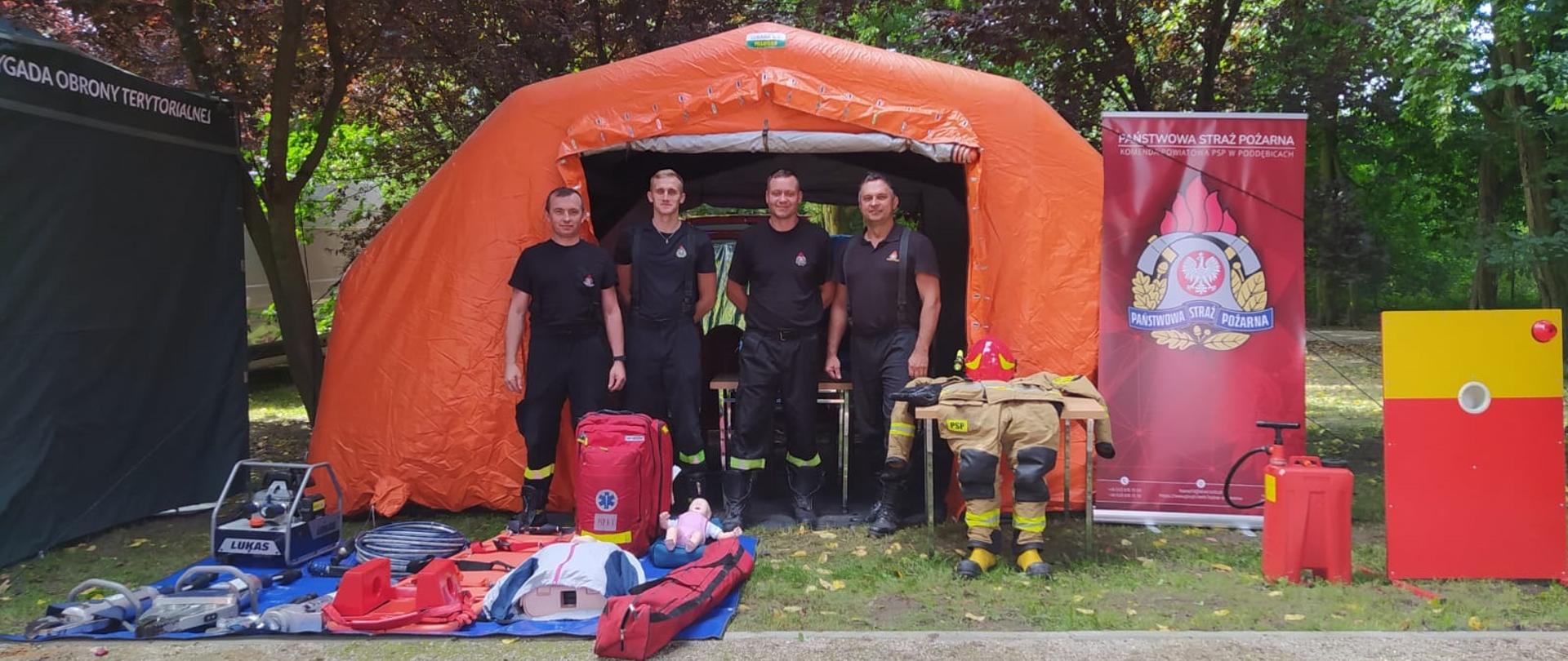 Czterej strażacy soją przed pomarańczowym dużym namiotem . Obok stoi baner z logo Państwowej Straży Pożarnej Przed namiotem na niebieskiej plandece rozłożone są różne drobne sprzęty używane przez strażaków w trakcie akcji ratowniczych Na stoliku leży strój strażacki Zdjęcie wykonane w plenerze za namiotem widać drzewa 