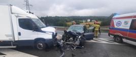 Zdjęcie ukazuje wypadek dwóch samochodów osobowego i dostawczego na drodze Krajowej nr 94. Jest pora dzienna. Po lewej stronie widzimy uszkodzony przód białego pojazd dostawczy. Obok tego pojazdu zjadają się zwrócony przodem pojazd osobowy z uszkodzonym cały przodem i otwartymi lewymi przednimi drzwiami. Za tym pojazdem osobowym stoi czterech strażacy ubranych w piaskowe ubrania i czerwonych hełmach. Po prawej stronie znajduje się część pojazdu Zespołu Ratownictwa Medycznego. Przed pojazdami znajdują się uszkodzone części pojazdów.