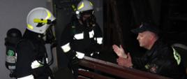 Widok z przodu. Po lewej stronie dwóch strażaków OSP w ubraniach specjalnych, hełmach i sprzęcie ochrony dróg oddechowych, po prawej stronie instruktor – oficer PSP - w trakcie zadania praktycznego.