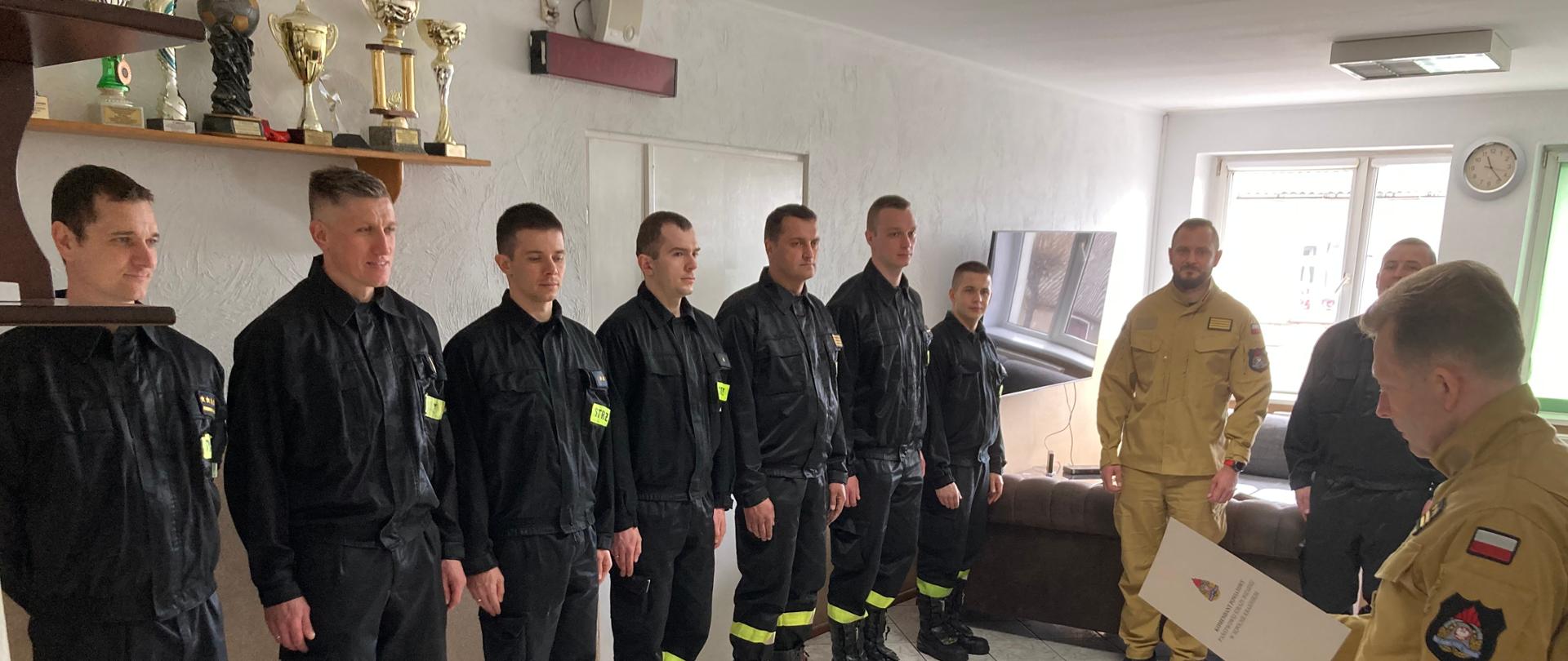 Zdjęcie wykonano wciągu dnia w budynku Komendy Powiatowej Państwowej Straży Pożarnej w Sępólnie Krajeńskim w pomieszczeniu świetlicy. Wzdłuż ściany stoją w szeregu ubrani w mundury koszarowe strażacy Państwowej Straży Pożarnej, naprzeciwko nich stoi Komendant Powiatowy ubrany w mundur służbowy trzymając w rękach pismo. 