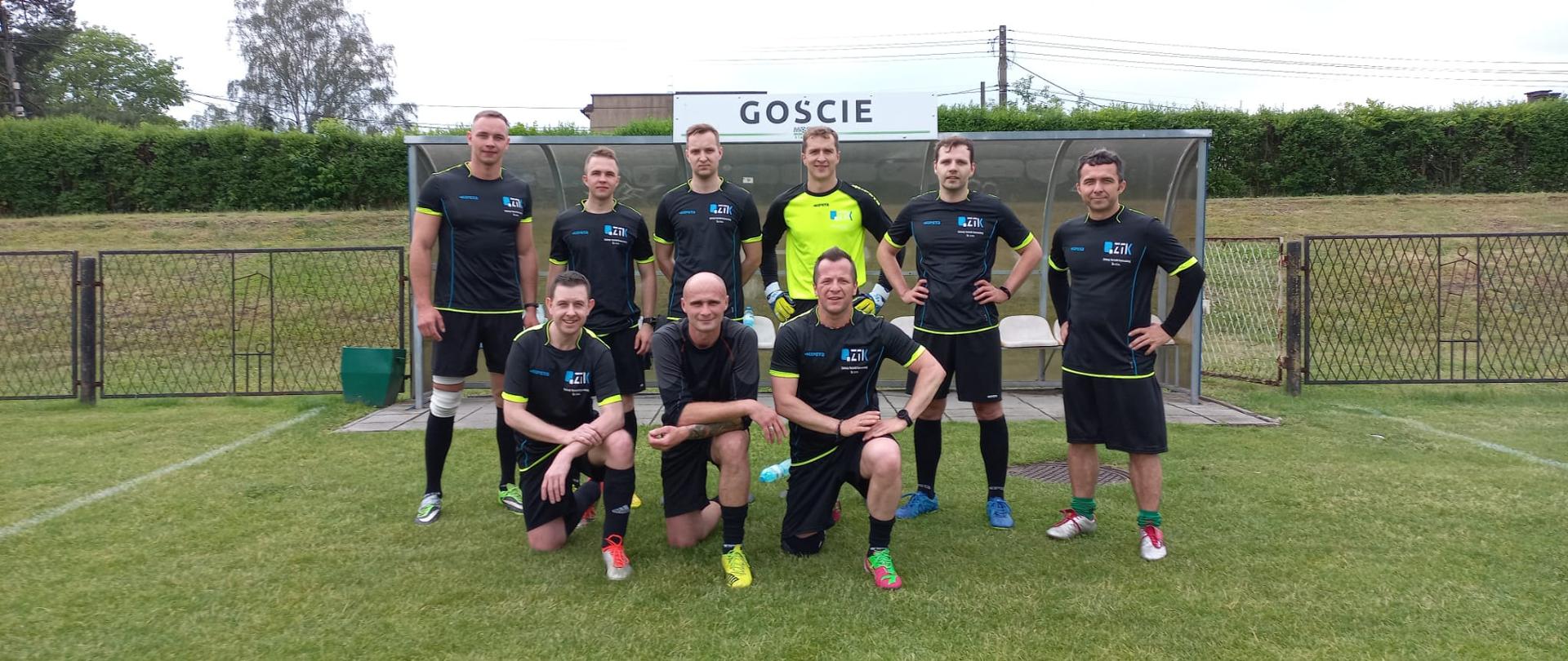Zawodnicy pozują do zdjęcia w bramce, pierwszy rząd klęczy, drugi stoi. Nad ich głowami napis GOŚCIE.
