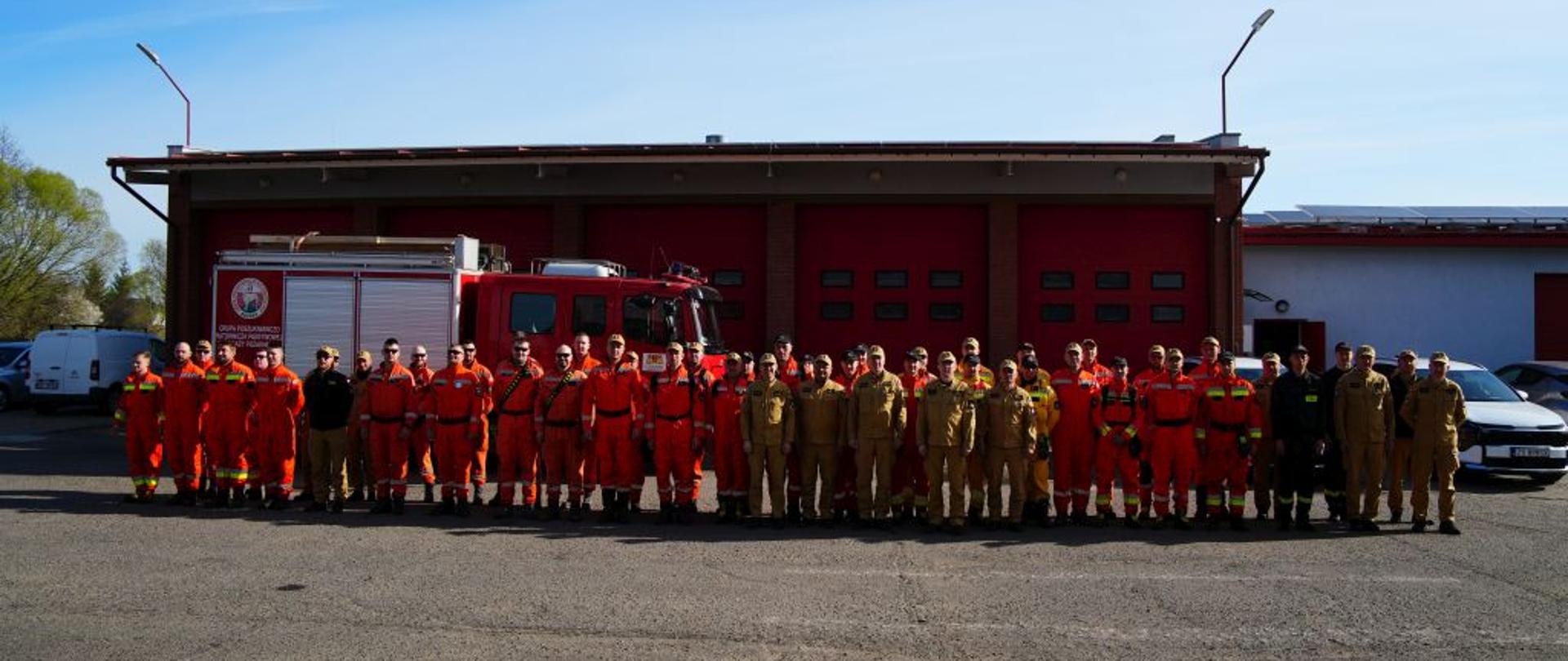 Zdjęcie grupowe dużej jednostki straży pożarnej na placu przed remizą. Kilkudziesięciu strażaków w pomarańczowych kombinezonach i piaskowych mundurach służbowych stoi w długim szeregu, twarzą do obiektywu. Za nimi zaparkowany jest duży, czerwony wóz strażacki oraz biały samochód osobowy. Nad budynkiem widać czyste, błękitne niebo. Zdjęcie wykonane jest z daleka, co pozwala na objęcie całej grupy i budynku strażnicy. Panuje słoneczna pogoda, a na placu widać długie cienie rzucane przez strażaków i budynek. Zdjęcie dokumentuje pamiątkową fotografię po zakończeniu ćwiczeń lub uroczystości.