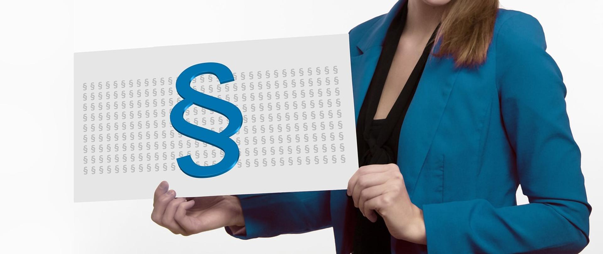 Pani w niebieskiej marynarce trzyma w ręku kartkę z niebieskim znakiem paragrafu. Nie widać jej twarzy. 