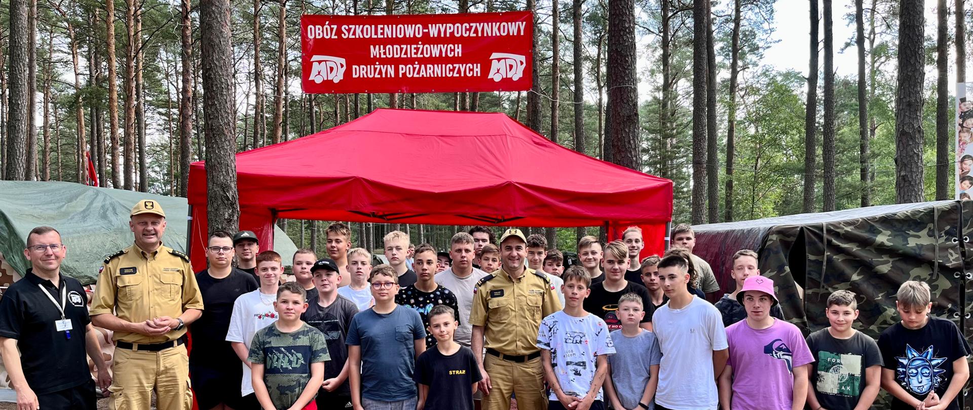 Pamiątkowe zdjęcie uczestników obozu i wizytujących gości. W tle czerwony namiot i baner obozu szkoleniowego, las i namioty uczestników