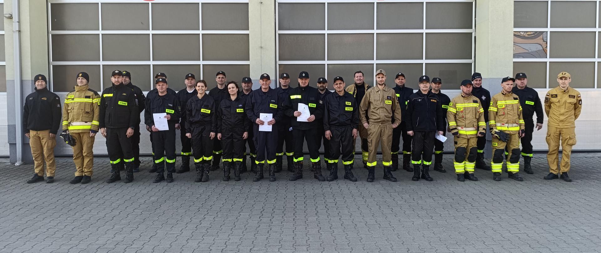 Zdjęcie przedstawia dużą grupę około 25-30 strażaków i strażaczek, stojących w kilku rzędach przed bramami garażowymi remizy strażackiej. Większość osób ubrana jest w ciemne, prawdopodobnie czarne, uniformy bojowe z żółtymi lub odblaskowymi elementami, oraz w jasnobeżowe, polowe mundury. Niektórzy trzymają w rękach białe dokumenty lub certyfikaty, co sugeruje zakończenie szkolenia lub inną formę uroczystości. W tle widoczne są trzy bramy garażowe z dużymi czerwonymi cyframi "3", "4" i "5" nad nimi. Nad bramami, na budynku, znajduje się napis "PAŃSTWOWA STRAŻ POŻARNA". Posadzka przed budynkiem jest wybrukowana szarymi kostkami. Atmosfera na zdjęciu jest uroczysta i formalna.