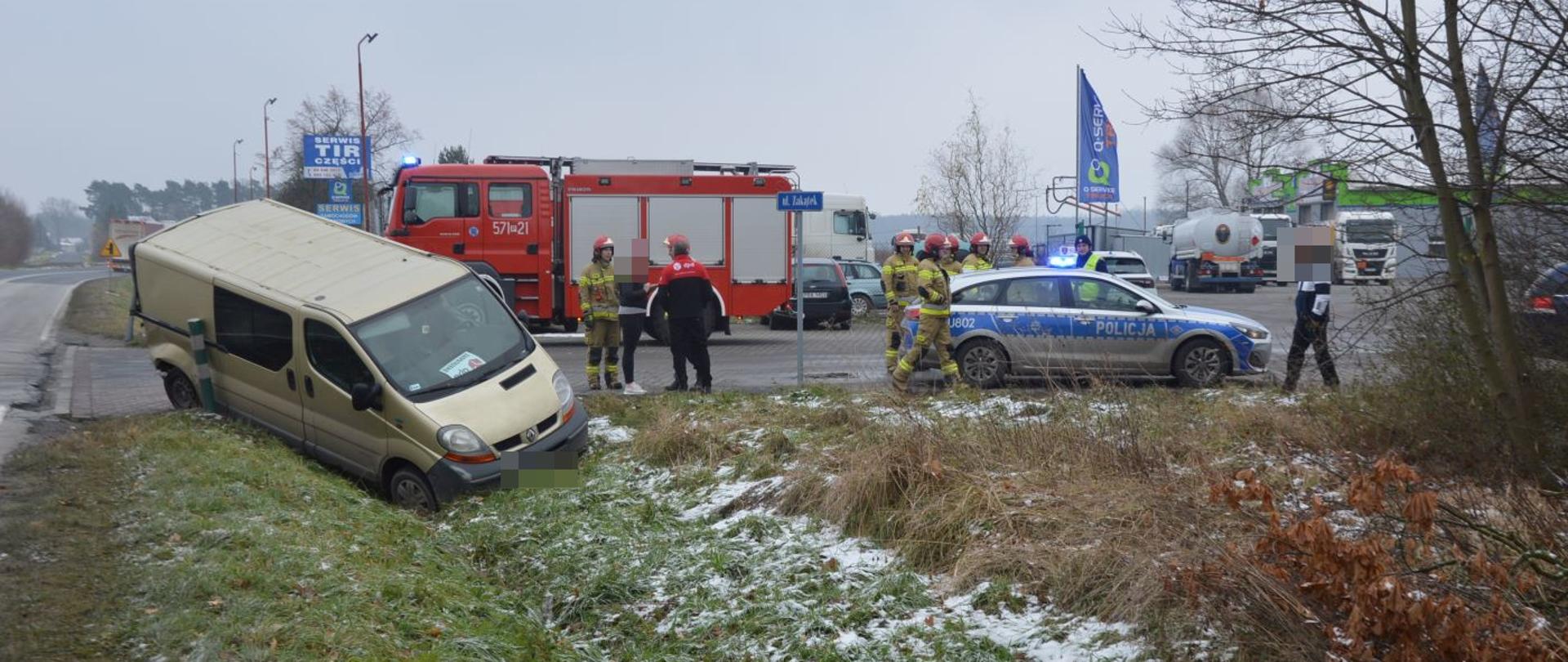 Kolizja dostawczaków. Droga gminna w Dębnie Polskim. Ogólny obraz miejsca kolizji - widok od strony Rawicza. Częściowo w rowie znajduje się renault trafic - jeden z pojazdów biorących udział w kolizji. Z prawej strony - radiowóz policyjny i wóz straży pożarnej. Obok samochodów strażacy, policjant oraz uczestnicy zdarzenia. W tle teren jednej z firm i pojazdy ciężarowe, w tym autocysterna.