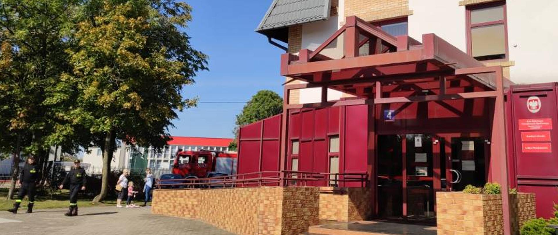 Zdjęcie przedstawia dwóch strażaków w ubraniach koszarowych podczas obserwacji próbnej ewakuacji prowadzonej w Placówce Terenowej Kasy Rolniczego Ubezpieczenia Społecznego w Zambrowie. Na zdjęciu widać część budynku Placówki, ewakuowanych interesantów oraz fragment wozu strażackiego. Zdjęcie w słoneczny dzień wykonano przed budynkiem Placówki Terenowej Kasy Rolniczego Ubezpieczenia Społecznego w Zambrowie. 