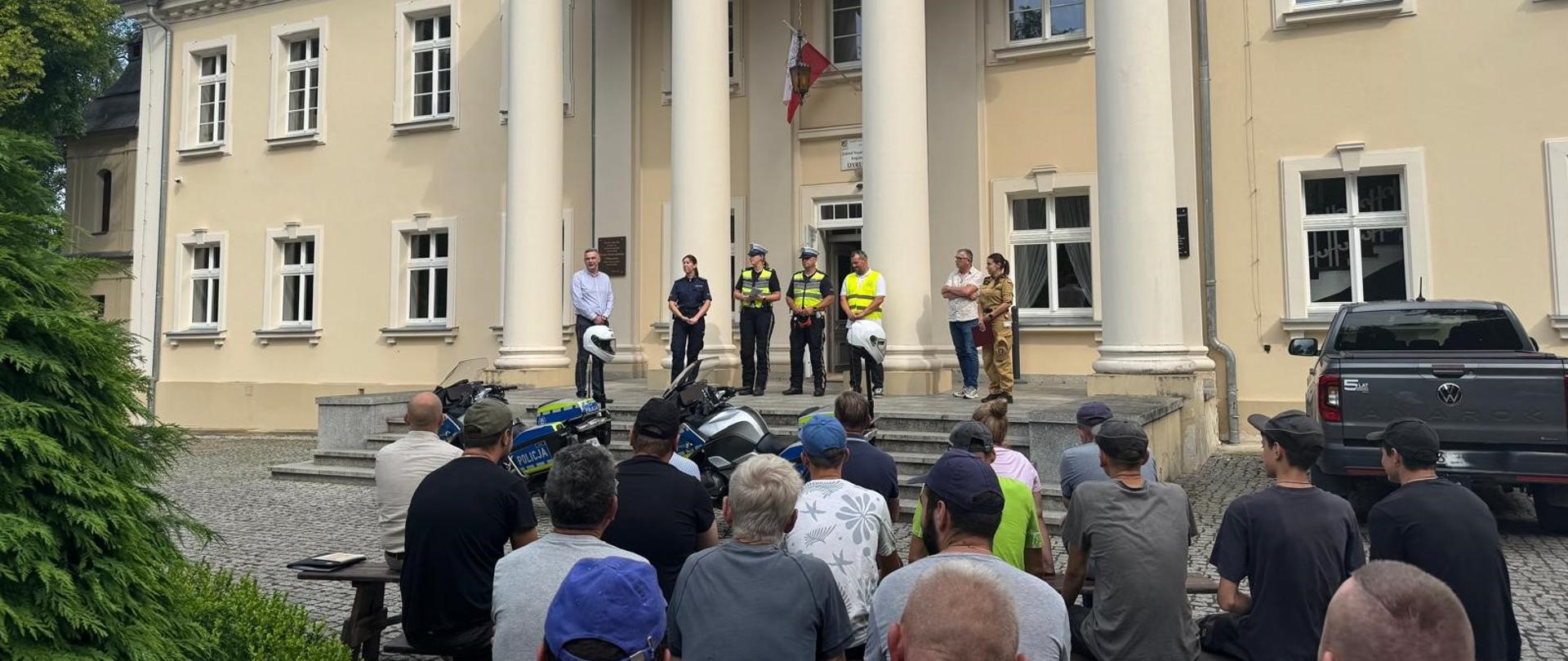 Na dziedzińcu przed jasnym, klasycystycznym budynkiem z czterema wysokimi kolumnami zgromadziła się grupa kilkudziesięciu osób siedzących na ławkach i słuchających wystąpień. Na schodach stoją przedstawiciele policji w umundurowaniu, osoby w odzieży odblaskowej oraz przedstawiciele władz lokalnych. Przed schodami ustawiono policyjne motocykle, a nad wejściem powiewa flaga Polski.
