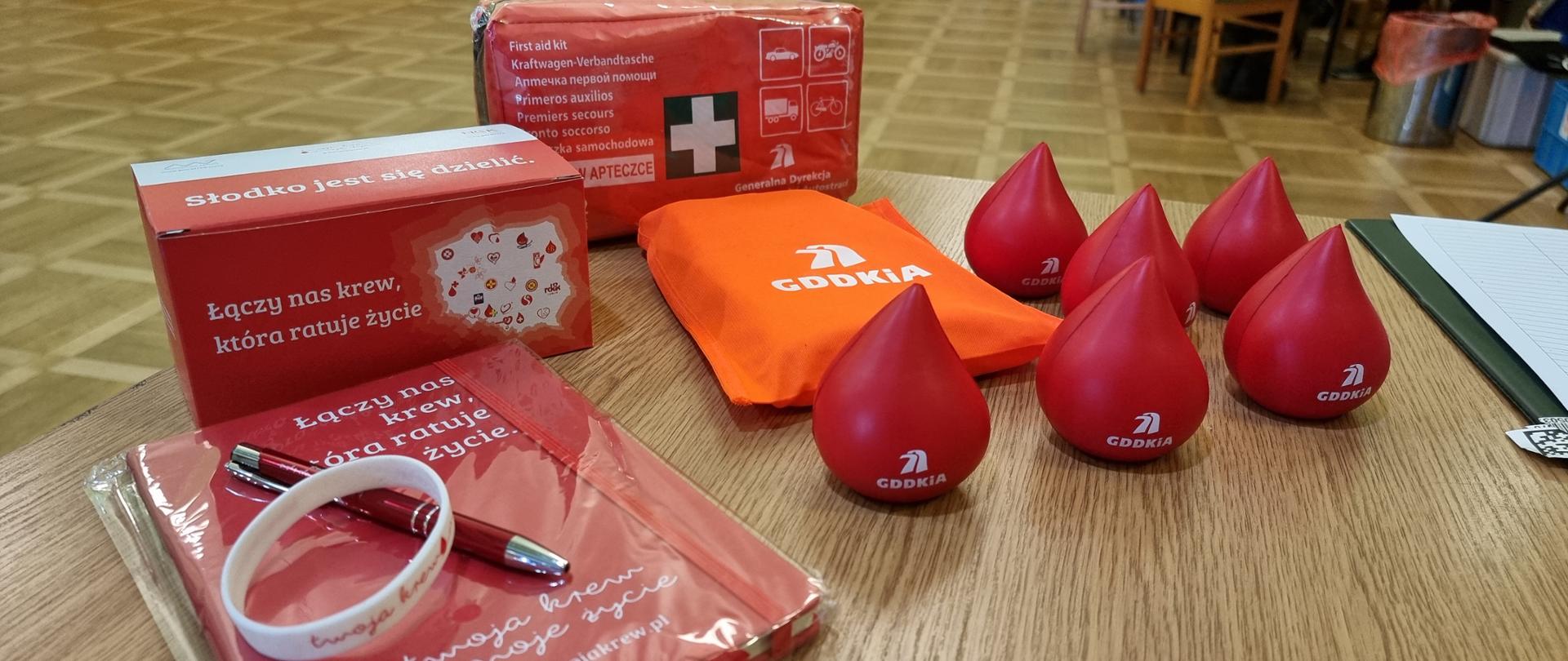 na zdjęciu widoczny stół na którym ułożone zostały gniotki w kształcie kropli krwi, apteczki, długopis i kamizelka z logo GDDKiA,. W tle widać osoby oczekujące na donację