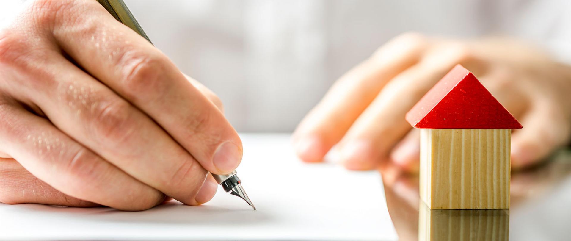 Dłoń trzymająca pióro, po prawej model domku.