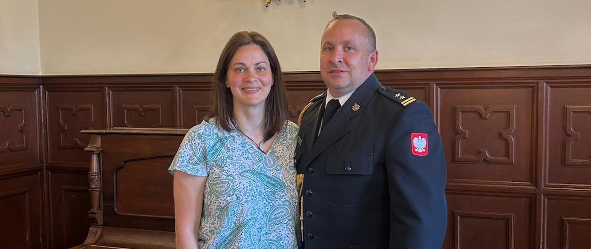 Na zdjęciu przedstawieni są Komendant Powiatowy Państwowej Straży Pożarnej w Pucku Pan bryg. Wojciech Wanat oraz mł. kpt. Grażyna Kwiek. oboje uśmiechają się do wykonywanego zdjęcia. Komendant jest ubrany w mundur galowy ze złotym sznurem na prawym ramieniu, natomiast Pani Grażyna Kwiek w ubranie cywilne.