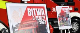 Na tle czerwonych samochodów strażackich stoją na sztaludze dwa plakaty na których widnieje napis "Bitwa o remizy 2023"