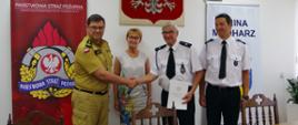 Widok z przodu. Uczestnicy spotkania stoją za stołem na tle rolapów KP PSP Wadowice i Gminy Mucharz. Od lewej: komendant powiatowy PSP, który ściska dłoń prezesa OSP, wójt gminy i naczelnik OSP.