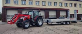 Czerwony ciągnik Massey Ferguson 5M z ładowaczem czołowym i osprzętem, zaparkowany przed budynkiem Komendy Powiatowej Państwowej Straży Pożarnej; do ciągnika podłączona jest dwuosiowa przyczepa transportowa Humbaur typu niskopodwoziowego.