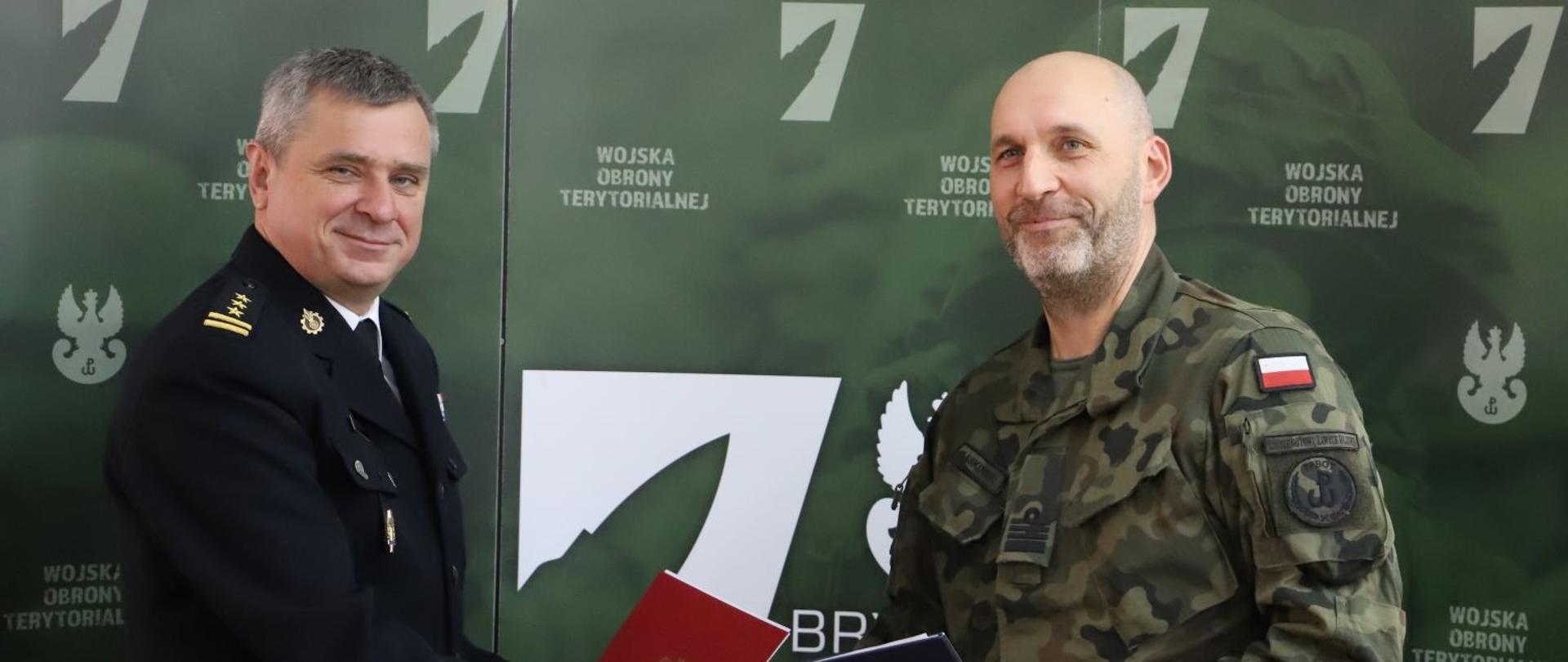 Pomorski komendant wojewódzki Państwowej Straży Pożarnej uściska dłoń dowódcy 7 Pomorskiej Brygady Obrony Terytorialnej po podpisaniu dokumentów. Obaj funkcjonariusze trzymają podpisane dokumenty.