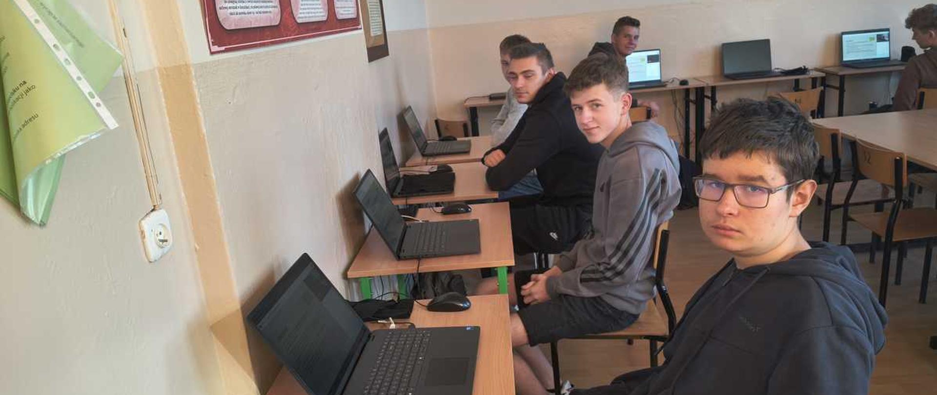 "Na zdjęciu widać salę komputerową w szkole, w której kilku nastoletnich chłopców siedzi przy biurkach i korzysta z laptopów.
Oto szczegółowy opis elementów znajdujących się na fotografii:
• Pierwszy plan (po prawej stronie): Najbliżej obiektywu siedzi chłopak w okularach, ubrany w ciemną, rozpinaną bluzę z kapturem. Patrzy bezpośrednio w stronę aparatu. Siedzi bokiem do biurka, trzymając ręce w kieszeniach.
• Drugi plan i głębia sali:
• Wzdłuż lewej ściany ustawiony jest rząd jednoosobowych biurek szkolnych na zielonych, metalowych stelażach. Na każdym biurku stoi otwarty, czarny laptop oraz myszka komputerowa.
• Przy kolejnych biurkach siedzą dwaj inni chłopcy. Pierwszy z nich, w szarej bluzie z kapturem marki Adidas, jest odwrócony przodem do obiektywu i lekko się uśmiecha. Za nim siedzi kolejny uczeń w czarnej bluzie, patrzący w stronę laptopa.
• W tle, po prawej stronie sali, widać kolejne biurka, przy których siedzą inni uczniowie. Stanowiska te są wyposażone w monitory stacjonarne lub laptopy.
• Otoczenie i ściany:
• Ściany pomieszczenia są jasne. Po lewej stronie, tuż przy krawędzi zdjęcia, wisi zielona tablica lub plakat informacyjny (widoczny jest fragment napisu „Ctrl+X”).
• Dalej na lewej ścianie wiszą czerwono-białe tablice edukacyjne lub dyplomy w ramkach.
• Na ścianie w głębi sali wiszą duże tablice korkowe z przypiętymi materiałami dydaktycznymi i infografikami.
• Oświetlenie i atmosfera: W sali panuje dzienne, naturalne oświetlenie, które wpada prawdopodobnie z okien niewidocznych na zdjęciu (po prawej stronie). Atmosfera wygląda na spokojną, typową dla lekcji informatyki lub zajęć dodatkowych."