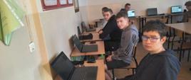 "Na zdjęciu widać salę komputerową w szkole, w której kilku nastoletnich chłopców siedzi przy biurkach i korzysta z laptopów.
Oto szczegółowy opis elementów znajdujących się na fotografii:
• Pierwszy plan (po prawej stronie): Najbliżej obiektywu siedzi chłopak w okularach, ubrany w ciemną, rozpinaną bluzę z kapturem. Patrzy bezpośrednio w stronę aparatu. Siedzi bokiem do biurka, trzymając ręce w kieszeniach.
• Drugi plan i głębia sali:
• Wzdłuż lewej ściany ustawiony jest rząd jednoosobowych biurek szkolnych na zielonych, metalowych stelażach. Na każdym biurku stoi otwarty, czarny laptop oraz myszka komputerowa.
• Przy kolejnych biurkach siedzą dwaj inni chłopcy. Pierwszy z nich, w szarej bluzie z kapturem marki Adidas, jest odwrócony przodem do obiektywu i lekko się uśmiecha. Za nim siedzi kolejny uczeń w czarnej bluzie, patrzący w stronę laptopa.
• W tle, po prawej stronie sali, widać kolejne biurka, przy których siedzą inni uczniowie. Stanowiska te są wyposażone w monitory stacjonarne lub laptopy.
• Otoczenie i ściany:
• Ściany pomieszczenia są jasne. Po lewej stronie, tuż przy krawędzi zdjęcia, wisi zielona tablica lub plakat informacyjny (widoczny jest fragment napisu „Ctrl+X”).
• Dalej na lewej ścianie wiszą czerwono-białe tablice edukacyjne lub dyplomy w ramkach.
• Na ścianie w głębi sali wiszą duże tablice korkowe z przypiętymi materiałami dydaktycznymi i infografikami.
• Oświetlenie i atmosfera: W sali panuje dzienne, naturalne oświetlenie, które wpada prawdopodobnie z okien niewidocznych na zdjęciu (po prawej stronie). Atmosfera wygląda na spokojną, typową dla lekcji informatyki lub zajęć dodatkowych."