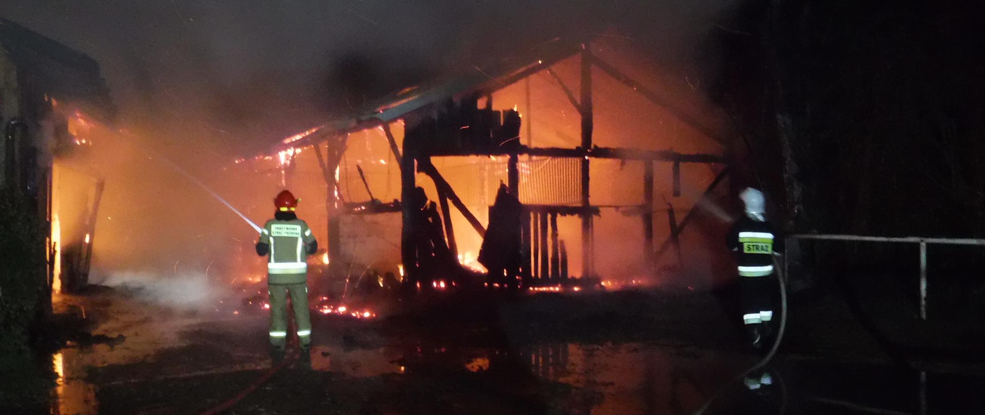 Zdjęcie przedstawia dwa drewniane budynki stajni w którym ma miejsce pożar. W budynkach widać ogień. Z dachu budynków widać unoszący się biały dym. Przed budynkiem stoją strażacy, po lewej i prawej stronie podając prądy wody na palący się budynek. Strażak po lewej ubrany w piaskowe ubranie specjalne i czerwony hełm. Strażak po prawej ubrany w czarne ubranie specjalne i biały hełm