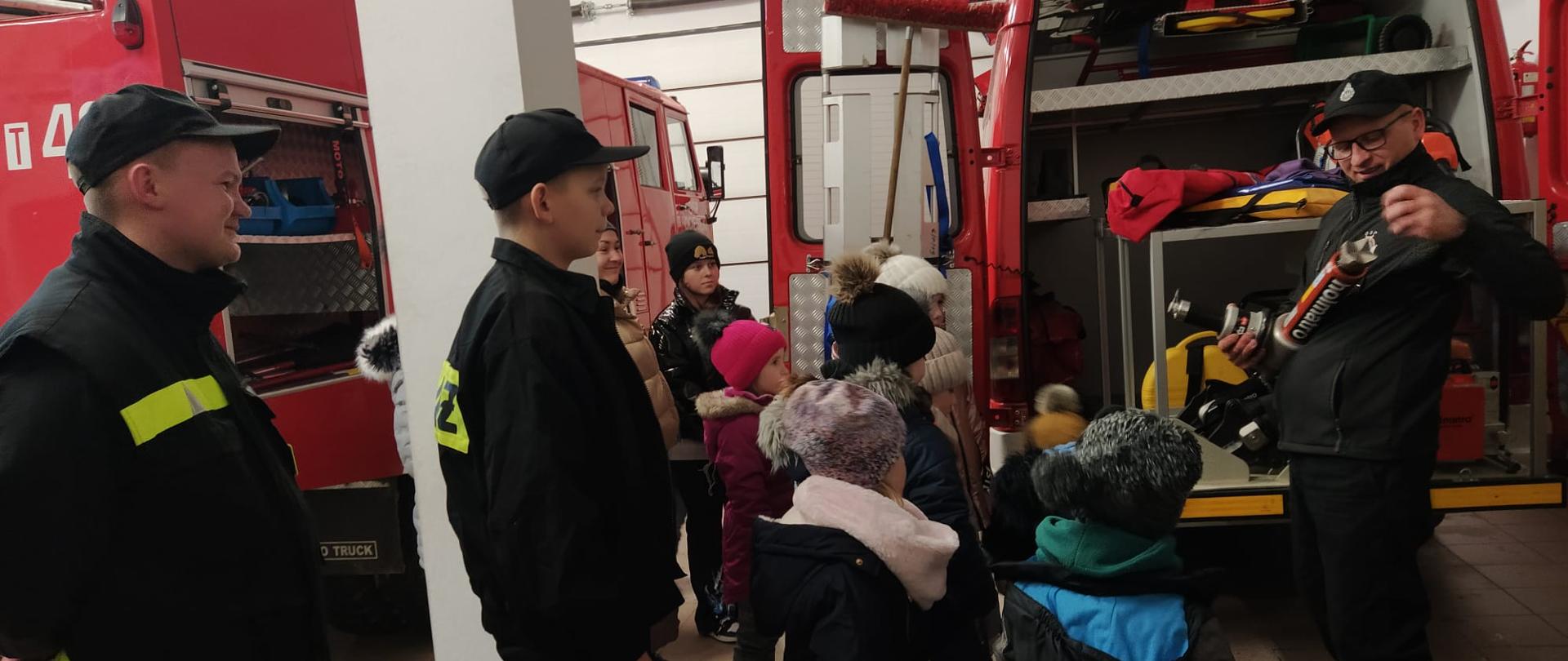 Zdjęcie prezentuje sprzęt pożarniczy. Dzieci oglądają wyposażenie wozu strażackiego. Druh omawia pojedyncze urządzenia. 