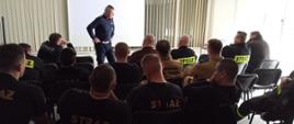 Zdjęcie przedstawia grupę strażaków oraz policjanta w sali wykładowej. Policjant prowadzi pogadankę dla strażaków.