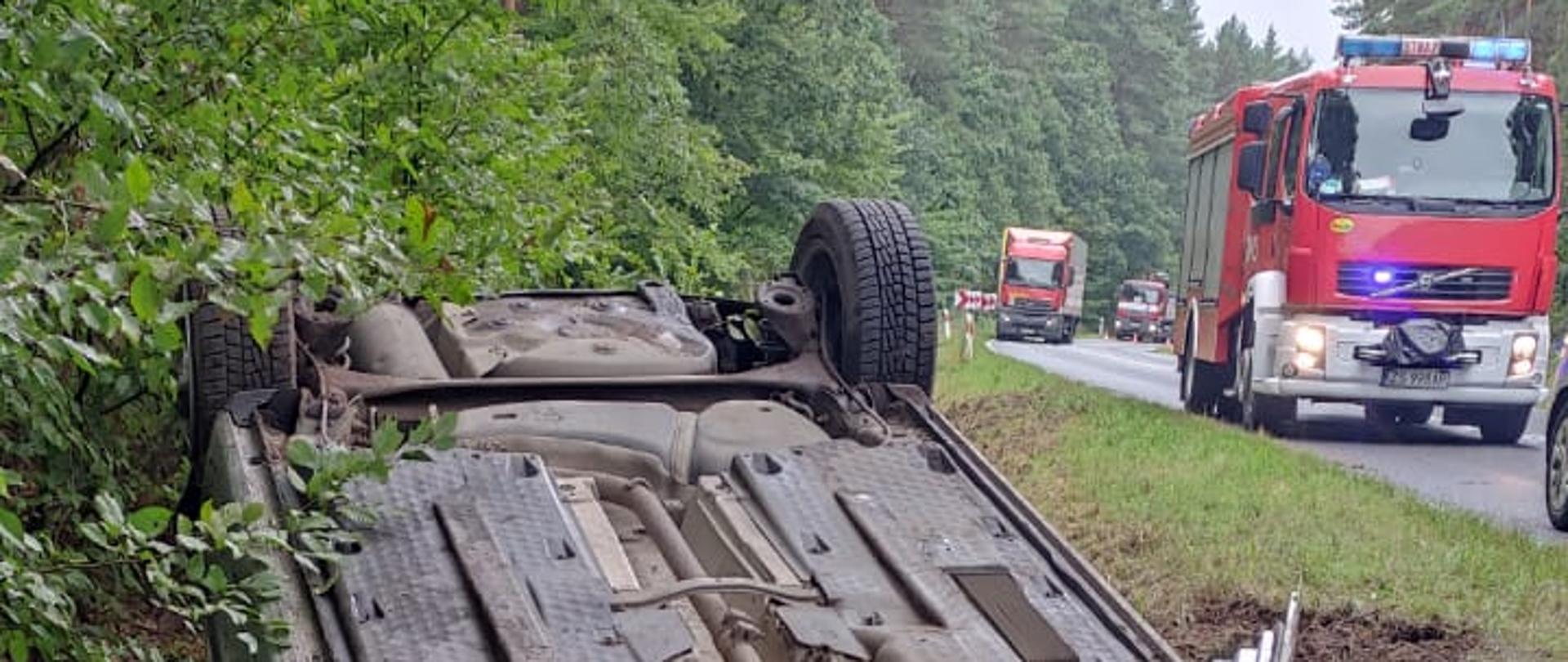 Na zdjęciu samochód osobowy leżący kołami do góry w rowie. Wokół las i jezdnia, na której widać także samochód pożarniczy.