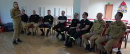 Świetlica komendy. Na krzesłach siedzą strażacy uczestniczący w zajęciach. Przed nimi stoi pani psycholog, która prowadzi psychoedukację. Wszyscy strażacy są umundurowani. W tle na ścianie widoczne jest logo PSP i drzwi ewakuacyjne. Z lewej strony, przy ścianie ustawione są stoły.
