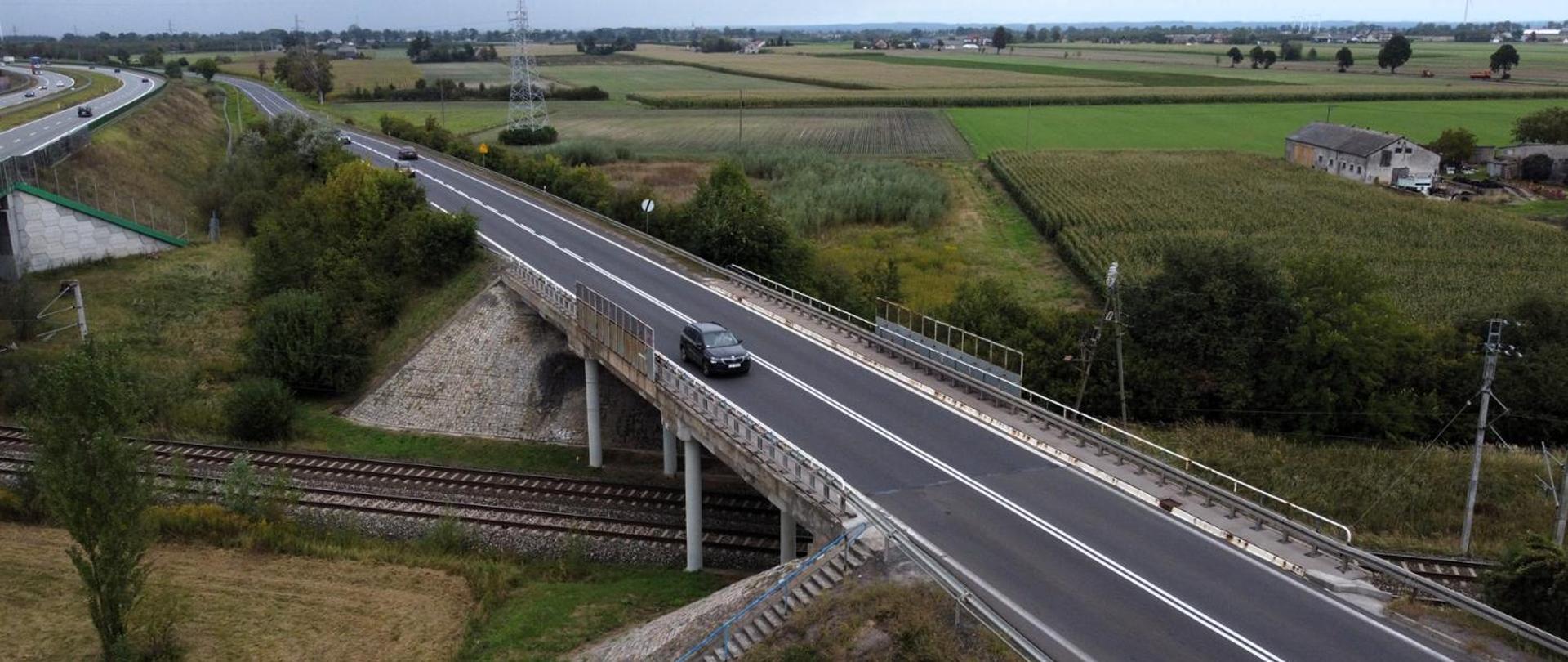 Widok z góry na drogę krajową przebiegającą nad torami kolejowymi, z jednym samochodem jadącym po wiadukcie. Po lewej stronie widoczna jest droga szybkiego ruchu, a w tle rozciągają się pola uprawne i zabudowania wiejskie.