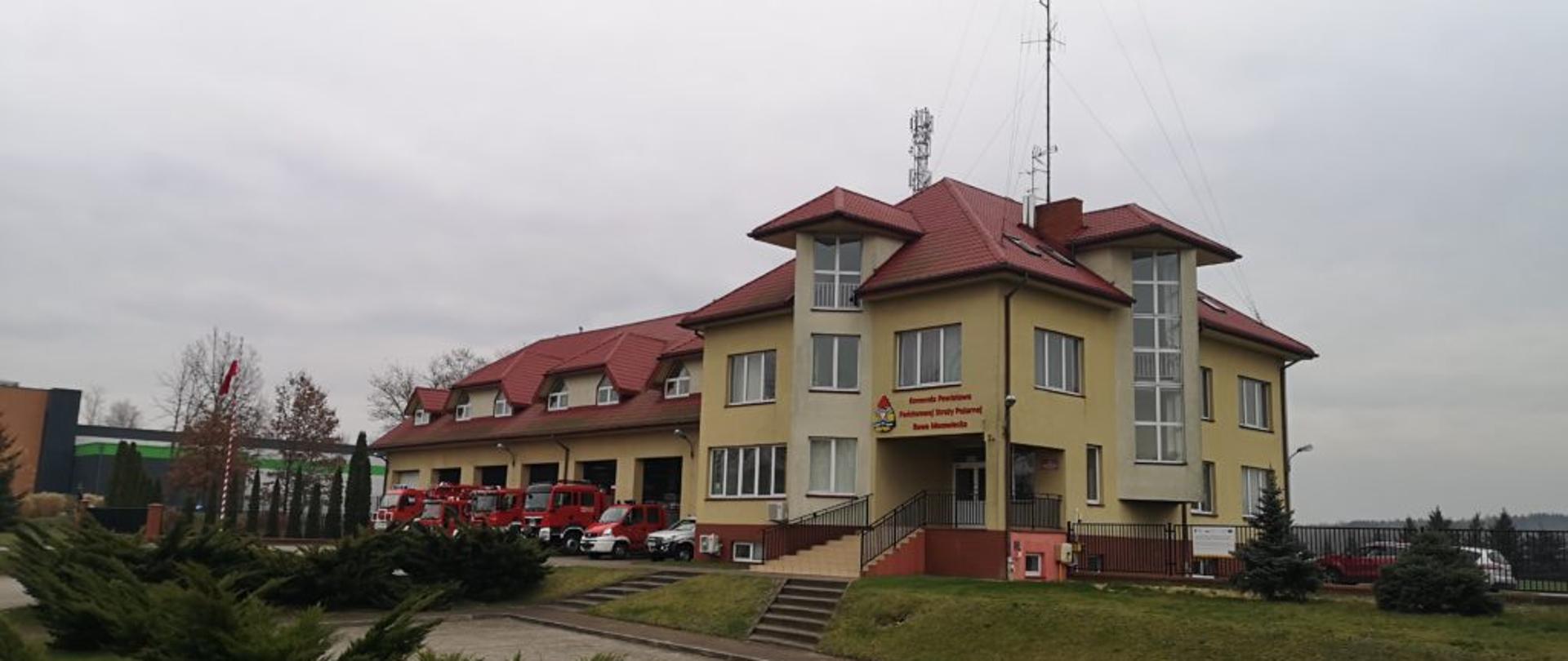 Zdjęcie przedstawia budynek Komendy Powiatowej Państwowej Straży Pożarnej w Rawie Mazowieckiej. Budynek znajduje się wyżej poziomu ulicy i parkingu dla interesantów. Budynek składa się z dwóch połączonych ze sobą części. Pierwsza, od lewej strony to część garażowa (parter + piętro), a druga to część administracyjna (piwnica + 3 piętra) Zdjęcie zrobione frontu. Budynek jest koloru żółtego i ma czerwoną dachówkę. Przed częścią garażową są wystawione samochody strażackie. 
