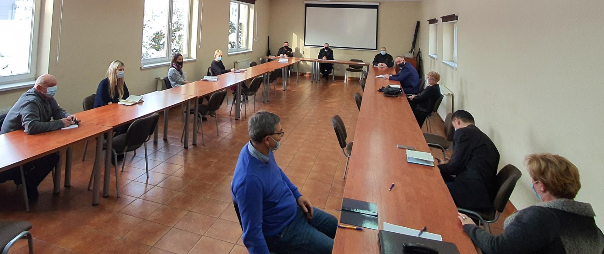 Zdjęcie wykonane w Sali Sztabowej Komendy Powiatowej Państwowej Straży Pożarnej w Będzinie podczas spotkanian dotyczącego programu szczepień. Na zdjęciu znajdująs się przedstawiciele samorządów terytorialnych z terenu powiatu będzińskiego oraz trzech funkcjonariuszy Komendy Powiatowej. Wszyscy uczestnicy spotkania siedzą przy stolikach.