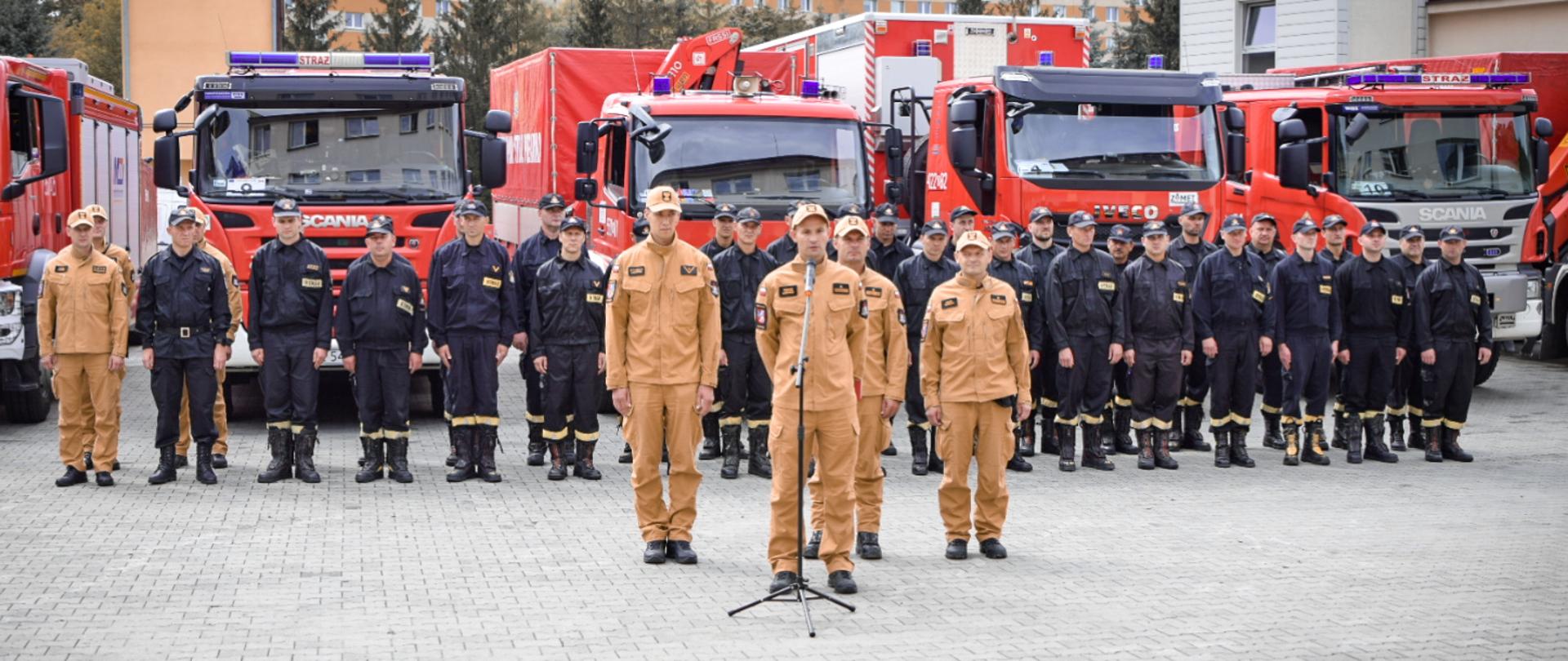 Zdjęcie zrobione na zewnątrz, podczas odprawy przed ćwiczeniami. W szeregu stoją ustawieni strażacy Państwowej Straży Pożarnej uczestniczący w ćwiczeniach. Przed mikrofonem stoi oficer Państwowej Straży Pożarnej w stopniu młodszego brygadiera. Za strażakami ustawione są samochody pożarnicze. 