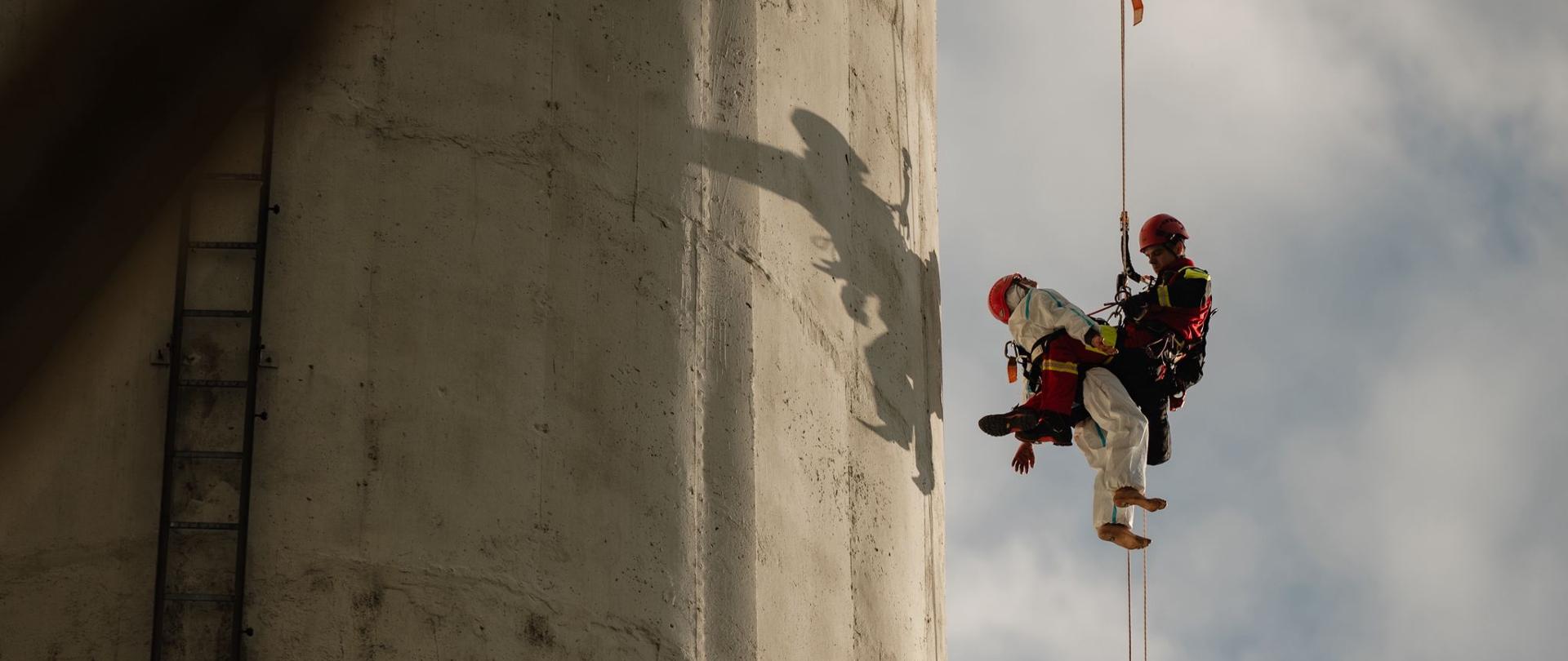 Ratownik wysokościowy PSP zjeżdża po linie z poszkodowanym w uprzęży z wysokości komina przemysłowego. W tle widoczny cień ratowników na betonowej konstrukcji.
