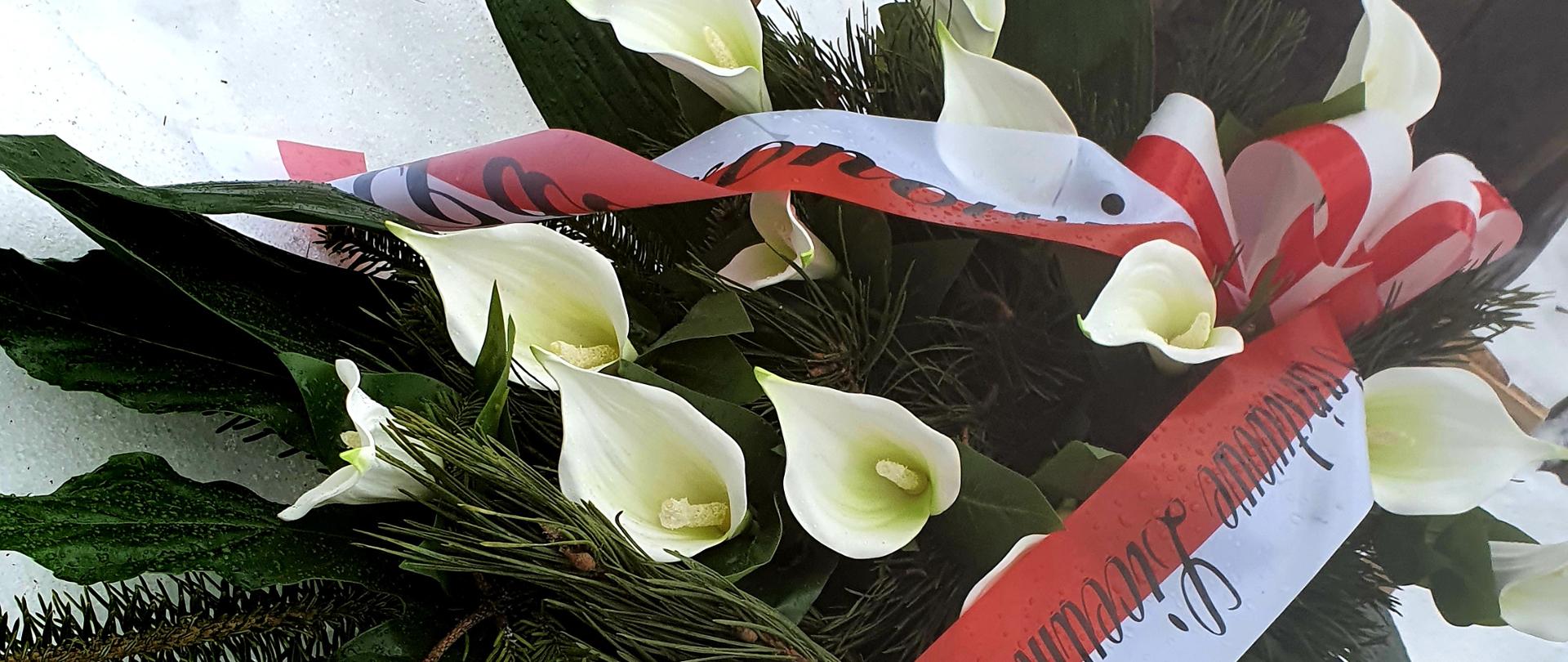 Na zdjęciu znajduje się wiązanka z białych lilii i zielonych liści i gałązek jodły. Wiązanka jest związana biało-czerwoną wstęgą z napisem: "Antoniemu Kenarowi - Patronowi Szkoły Państwowe Liceum Sztuk Plastycznych w Zakopanem". Wiązanka położona jest na śniegu, na grobie Antoniego Kenara.