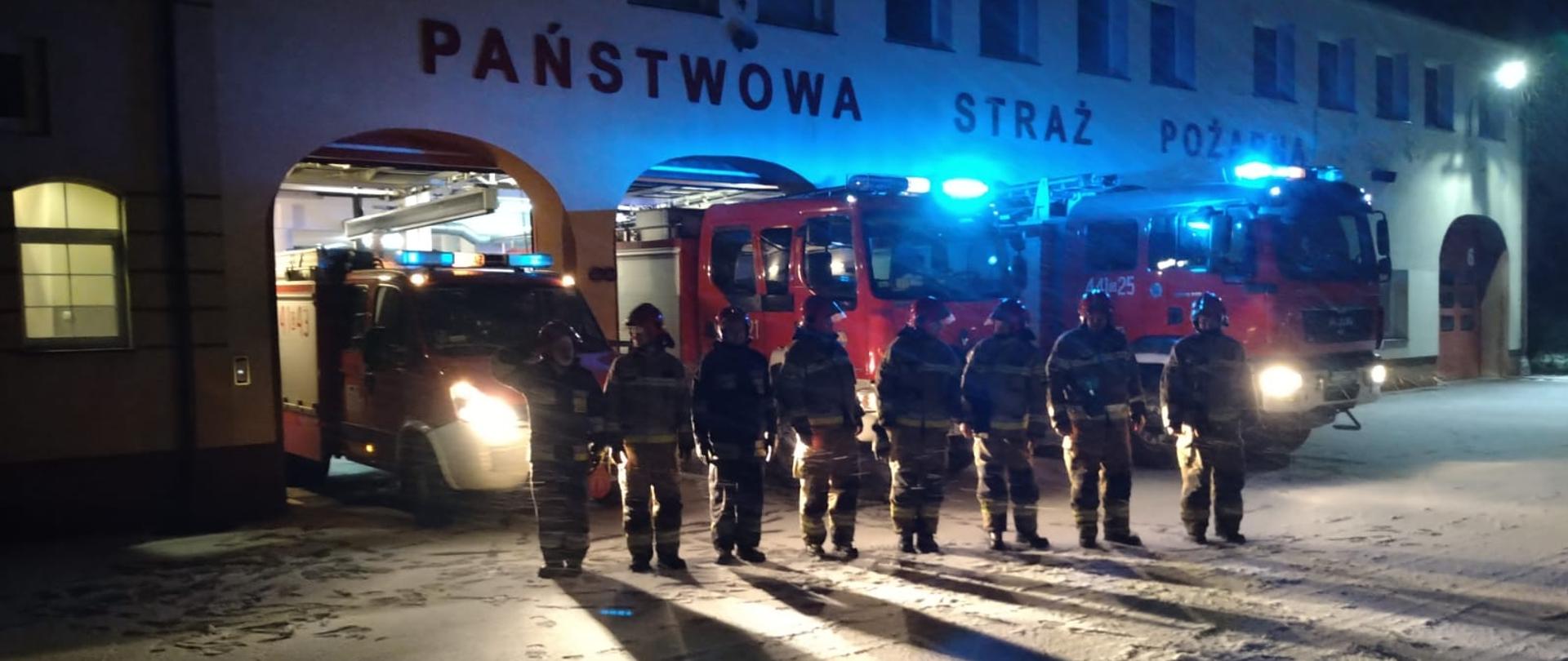 Strażacy ustawieni w szeregu na tle samochodów pożarniczych z załączoną sygnalizacją